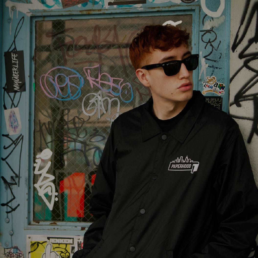 A man with short brown hair wearing black sunglasses and a black jacket with white Paperhood logo on the breast standing in front of blue door with a window that has been graffitied.