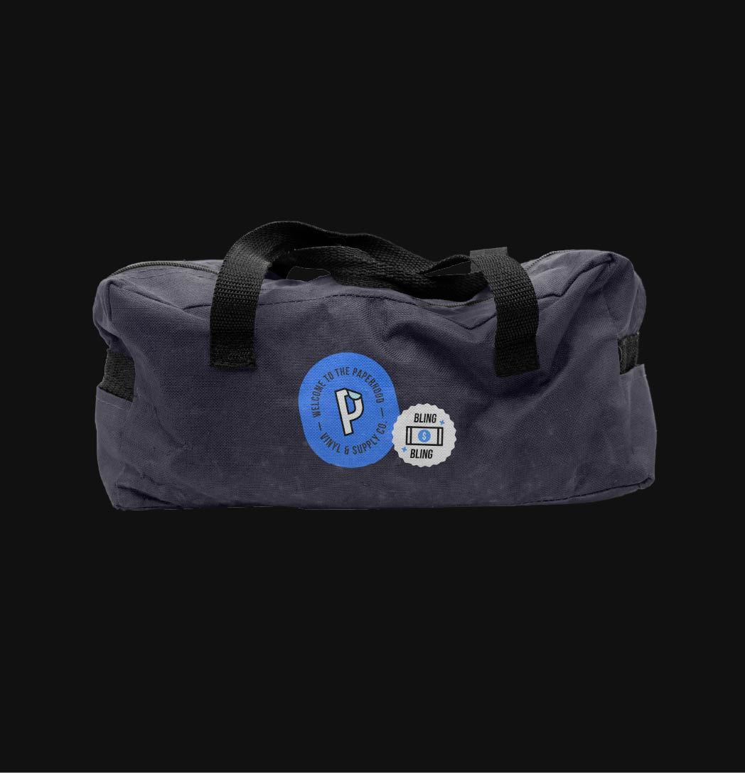 Dark blue-ish gray duffel bag with a black zipper and black handles with a large bright blue Paperhood logo patch and small white Paperhood icon patch over a black background.