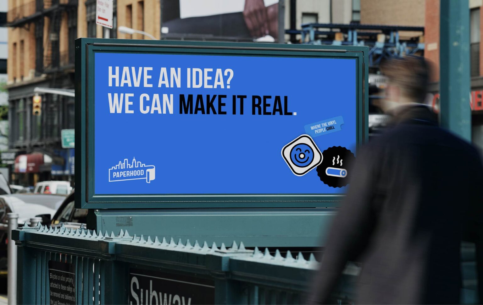 An outdoor billboard hung above a subway entrance in New York City with a person walking by, obscured by motion blur.