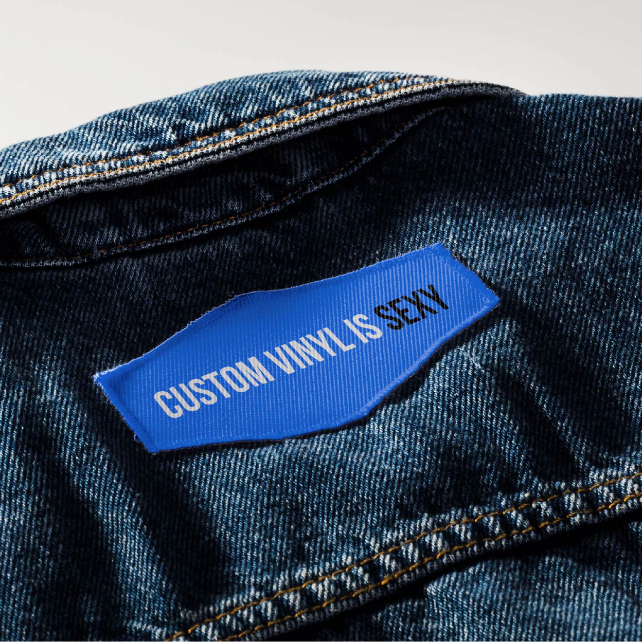Close up of a bright blue patch that says "Custom Vinyl is Sexy" on the back of a denim jacket, right below the jacket collar.