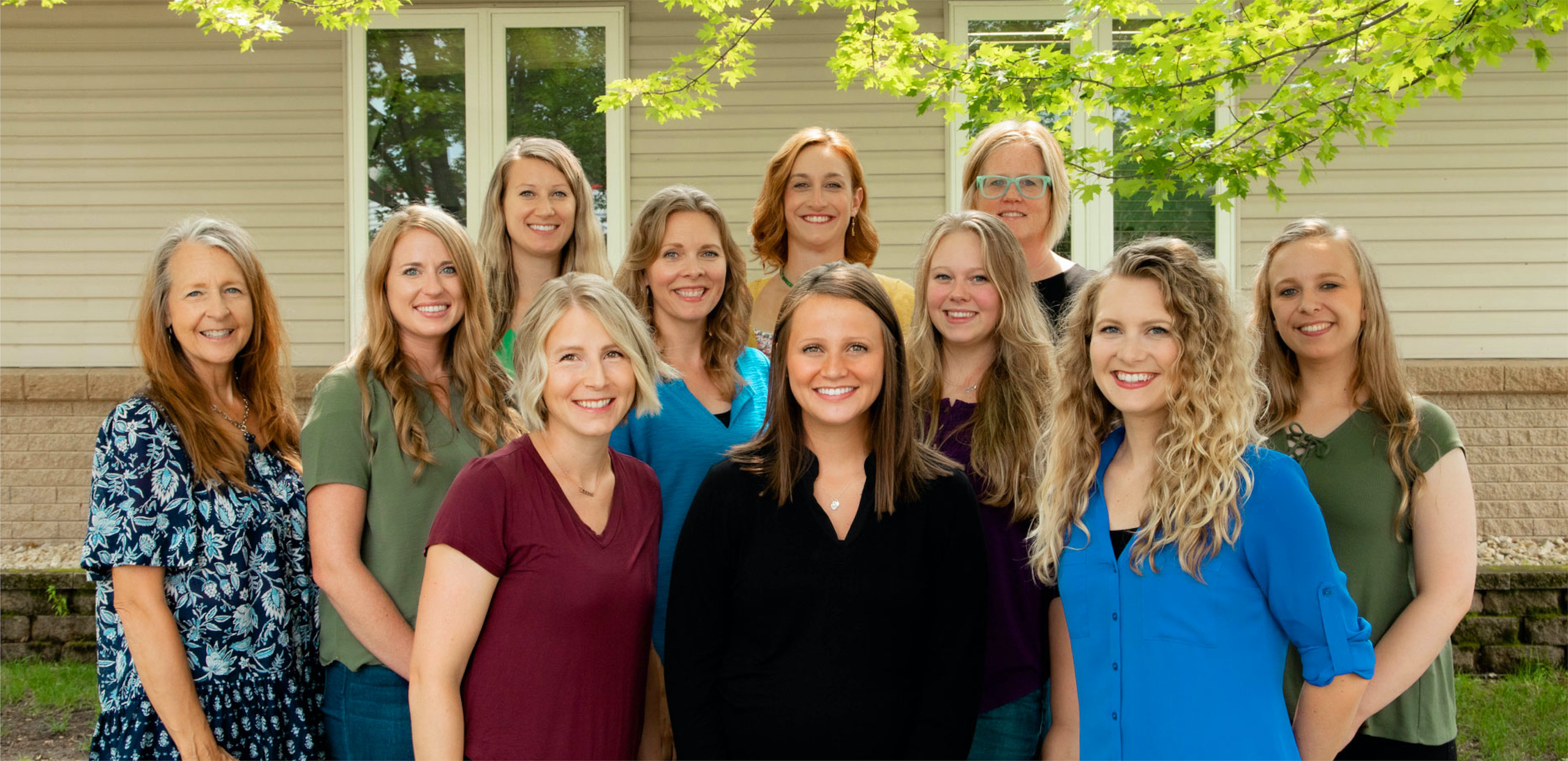 Group photo of 11 women smiling in front of a building with leafy tree branches peaking into frame.