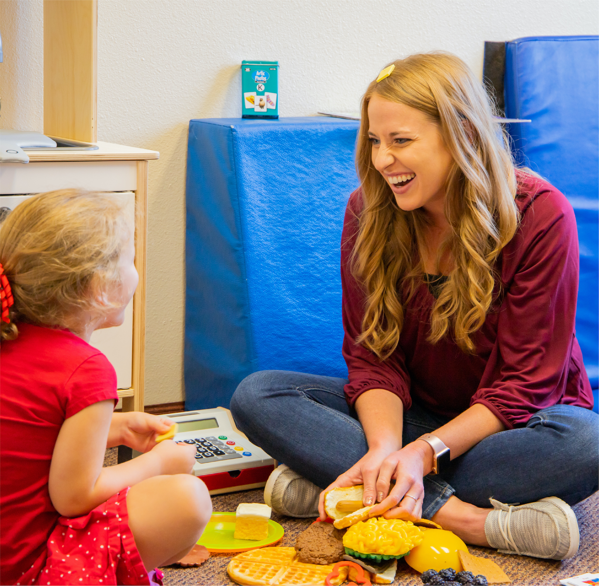 Blonde woman in a red shirt sitting cross-legged on the floor smiling at a little girl who is sitting cross-legged across from her, with toys on the floor in between them.