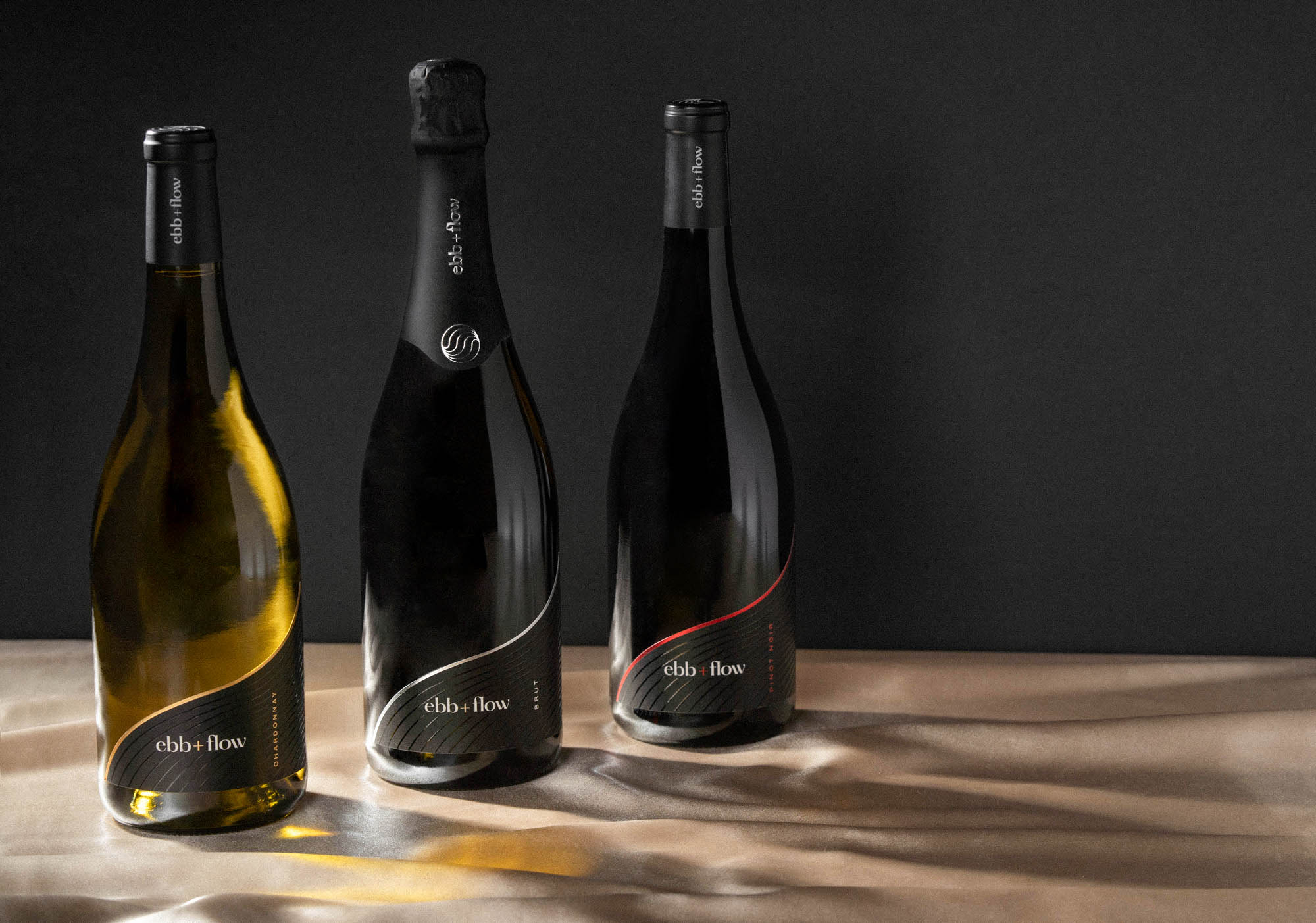 Three wine bottles in front of a black background and sitting on a light tan fabric. The labels read "ebb + flow" and each bottle has a different foil accent color - one is red, one is silver, and one is gold.