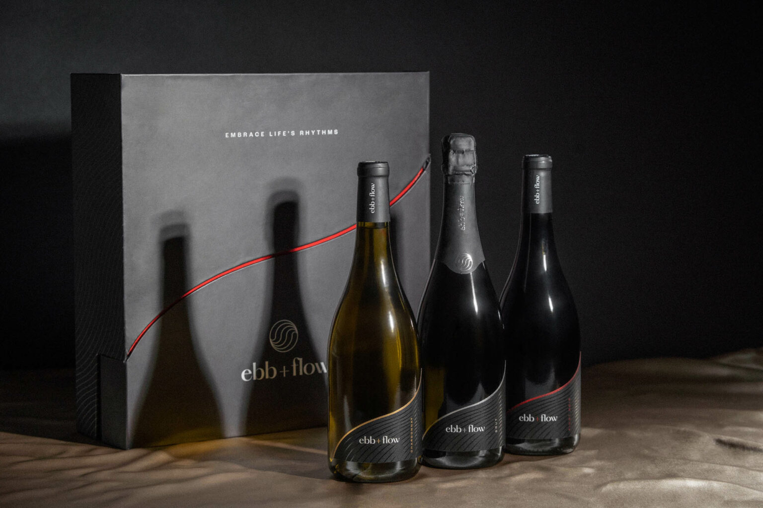 Sitting on a light tan fabric are three wine bottles in front of a black box. The labels read "ebb + flow" and each bottle has a different foil accent color - one is red, one is silver, and one is gold. The box has a red wavy line on the front, below that is the ebb + flow logo, and above it reads "Embrace Life's Rhythms"