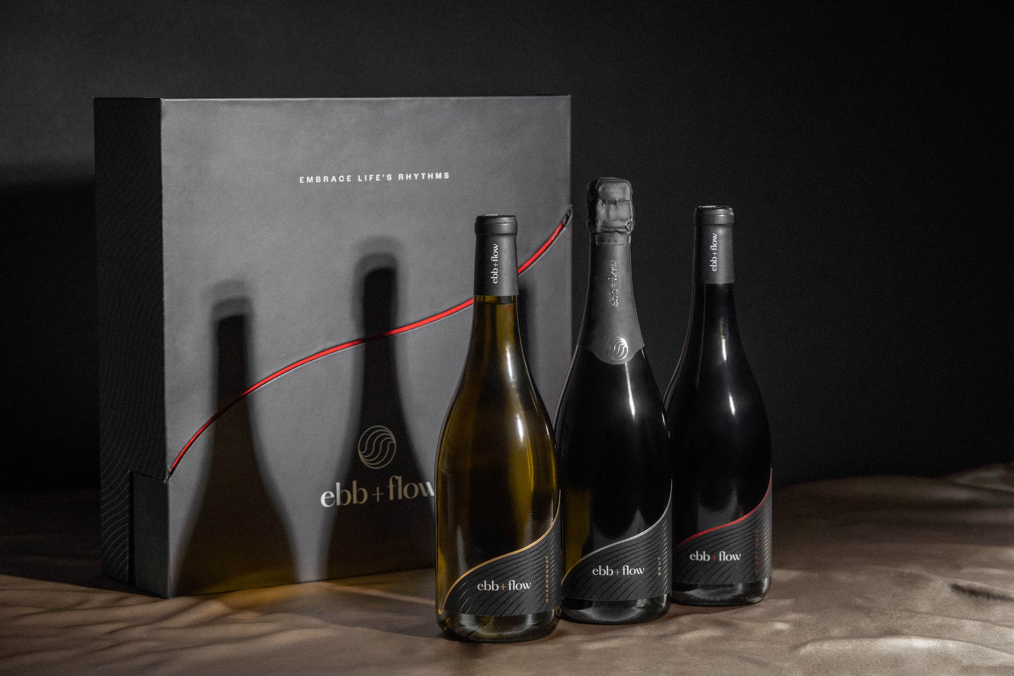 Sitting on a light tan fabric are three wine bottles in front of a black box. The labels read "ebb + flow" and each bottle has a different foil accent color - one is red, one is silver, and one is gold. The box has a red wavy line on the front, below that is the ebb + flow logo, and above it reads "Embrace Life's Rhythms"