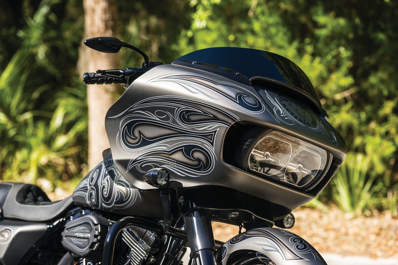 A close-up shot of a shiny grey motorcycle with decorative swashes on the front. In the background is blurred foliage and trees.