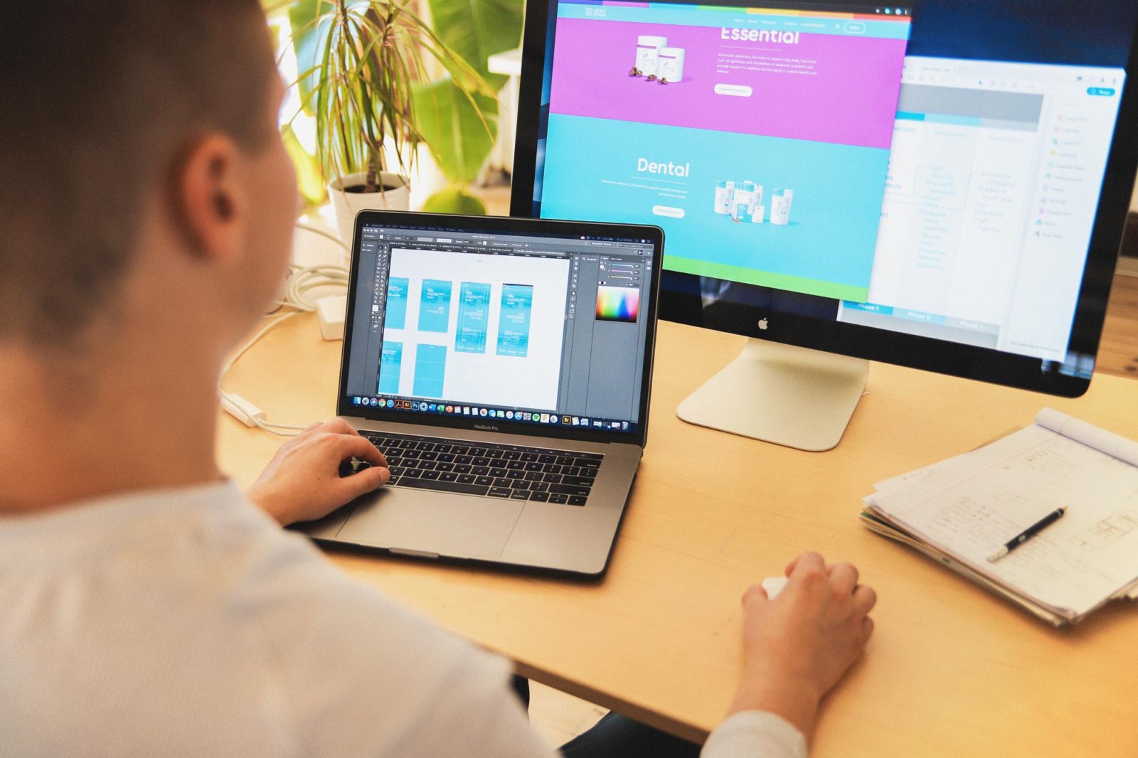 View peeking over a mans shoulder to show a browser window open on his monitor, he is using a mouse with his right hand and is using the keyboard on a laptop with his left hand.