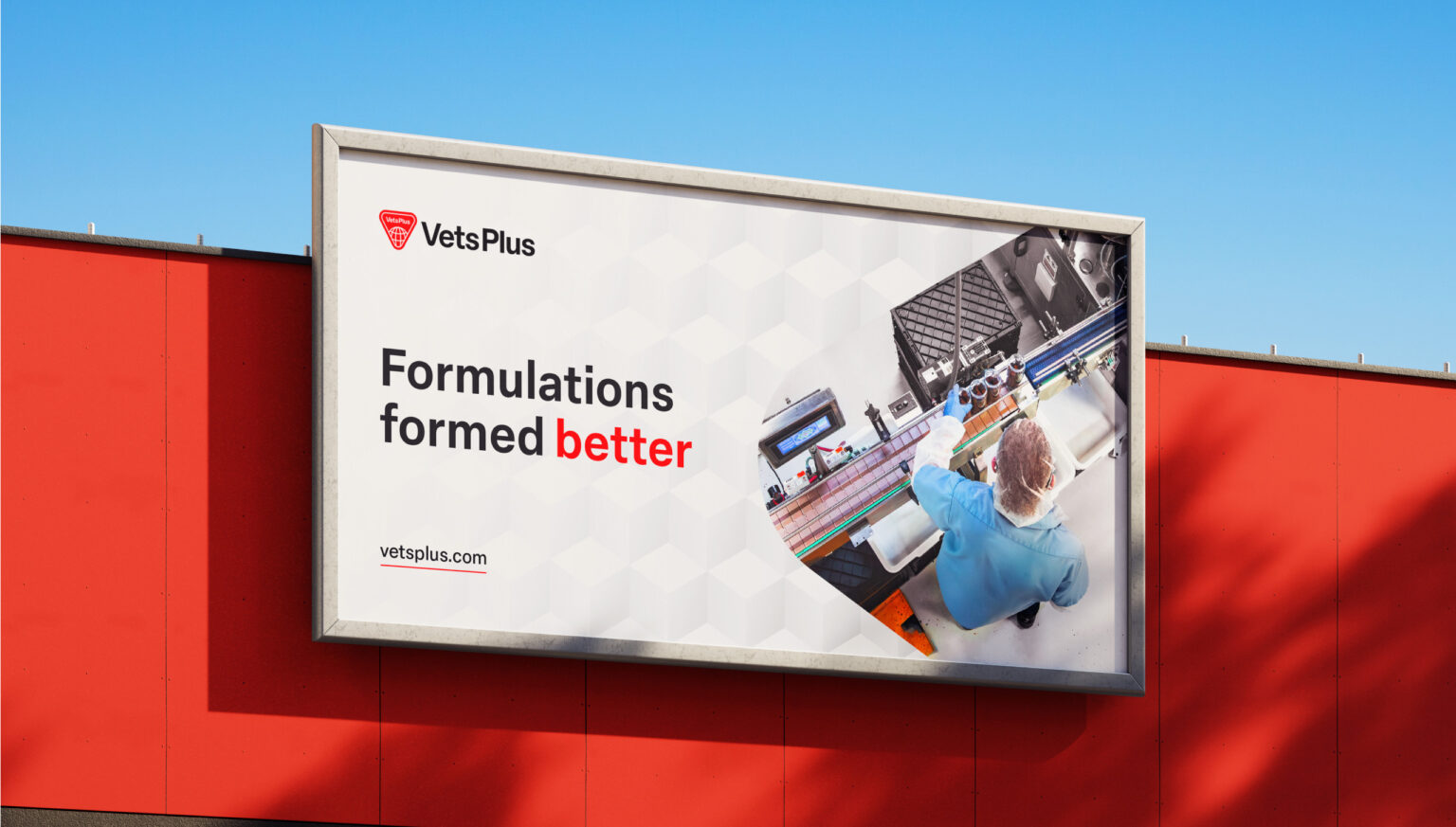 A billboard with a grey frame mounted on a bright red wall. On the lefthand side of the billboard is the VetsPlus logo, and below it reads "Formulations Formed Better". On the right hand side is an image of a person in a hairnet, blue shirt, and blue gloves operating a product line in a factory in a triangular holding shape.